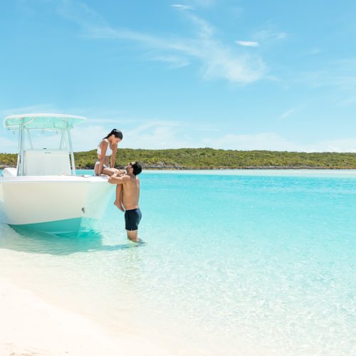 A man and woman in swimwear interact near a white boat anchored in shallow, clear blue water with a sandy beach and lush green coastline in the background under a bright, sunny sky.