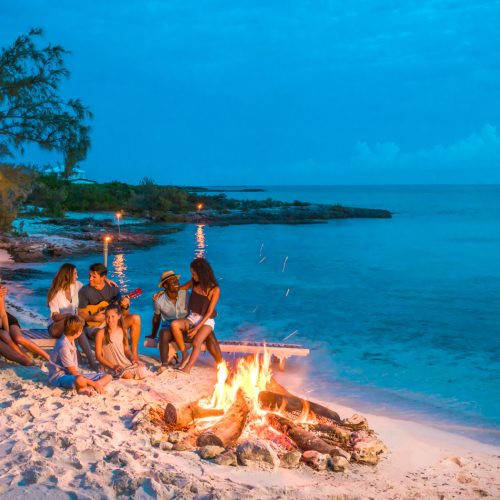 A group of people sitting around a bonfire on a beach at dusk. Some are playing guitars while others are talking and enjoying the evening. The sea is calm and the sky is turning dark blue.