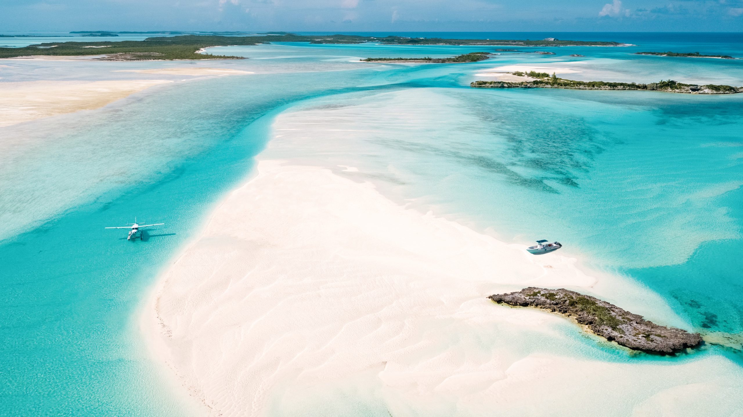 Aerial view of a sea plane and a boat on a sandbar surrounded by turquoise water, with islands in the distance.