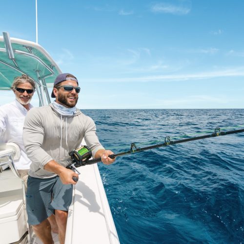 Two men stand on a boat in the ocean, one of them actively fishing with a rod while the other watches.