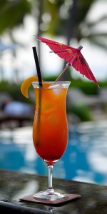 A tropical cocktail with a slice of orange, black straw, and red paper umbrella, sits on a coaster by a blurred poolside background.