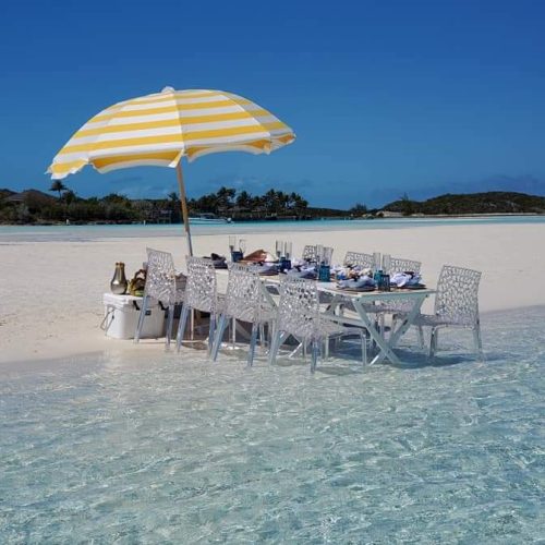 A dining table with wicker chairs is set up on a beach, under a yellow and white striped umbrella, with the clear, shallow water of the sea surrounding it.