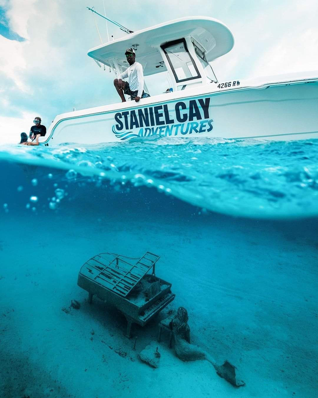 A boat with "Staniel Cay Adventures" on the side is on the water surface. Through the clear water, an underwater scene showing a submerged piano and a mermaid statue is visible.