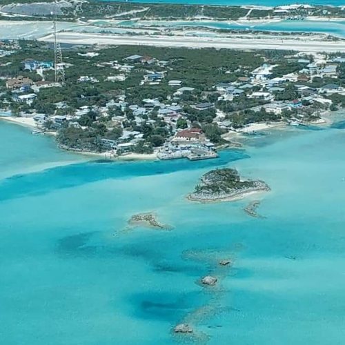 Aerial view of a coastal town surrounded by turquoise water and sandy beaches, with buildings scattered among greenery.