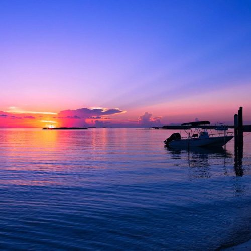 A small boat is moored at a dock on calm water during a vibrant sunset with an orange and purple sky.