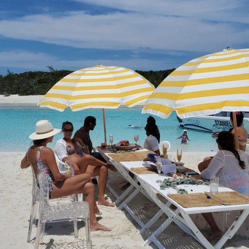 People sit and enjoy under yellow and white striped umbrellas by a beach, with a table set with food and drinks. The background shows clear turquoise water and a partially cloudy sky.