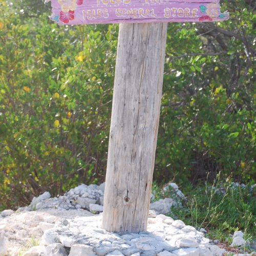 Weathered wooden signpost with a pink sign reading "Isles Inn" and "Isles General Store" to the right, surrounded by greenery and resting on a stone base.