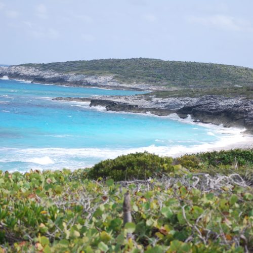 A coastal landscape with turquoise waters, white waves crashing on rocky shores, and greenery in the foreground under a partly cloudy sky.