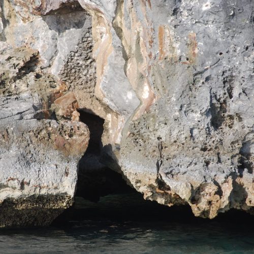 Close-up of a rocky cliff face with visible layers and color variations, featuring a small, dark cave opening at the base near the water.