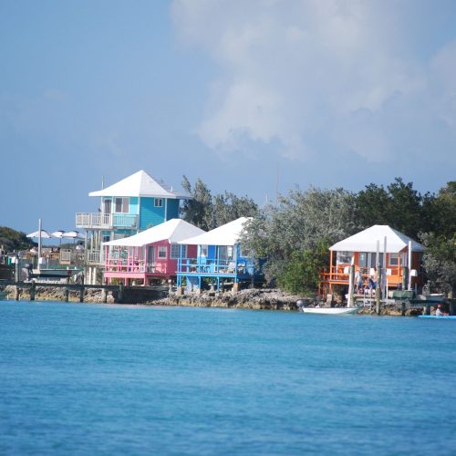 A row of colorful beach houses with white roofs line the shoreline against a backdrop of trees and blue sky, fronted by a calm turquoise sea.