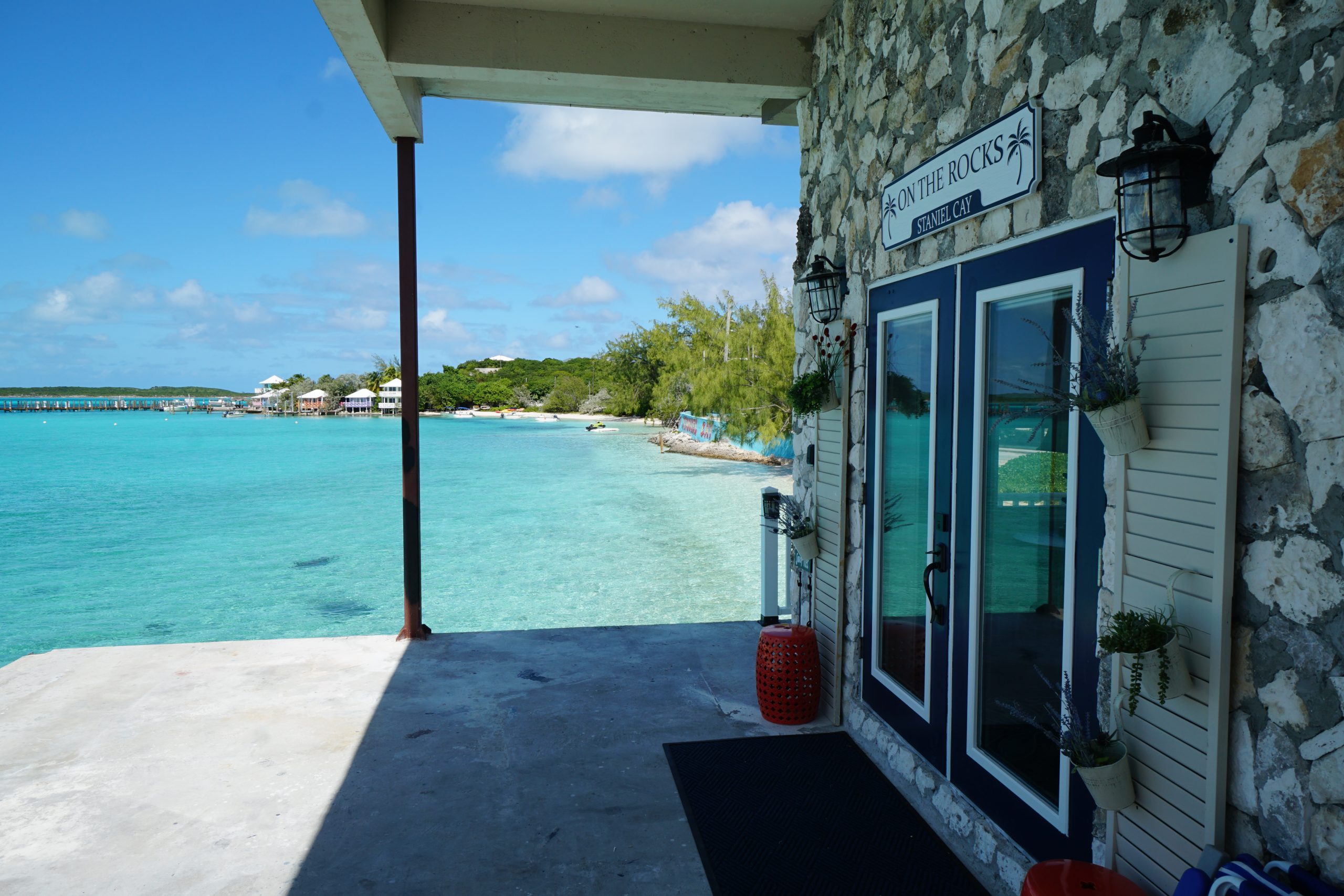 A stone building with a sign "On the Rocks" overlooks a clear turquoise sea, under a partly cloudy blue sky.