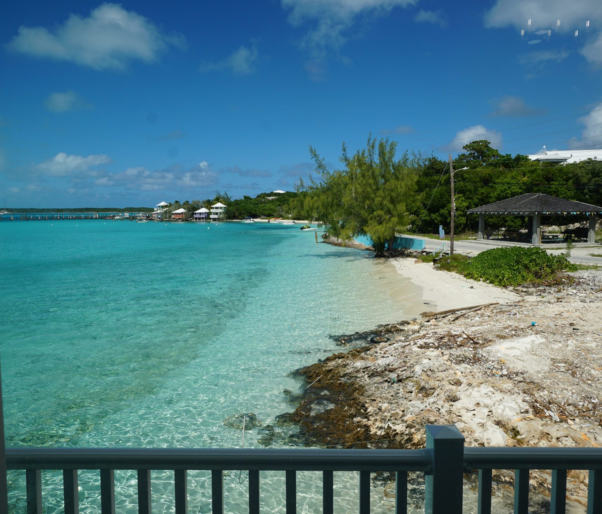 staniel Cay vacation Rentals View of a clear turquoise shoreline with a small beach, trees, and a pavilion on the right under a blue sky with scattered clouds.