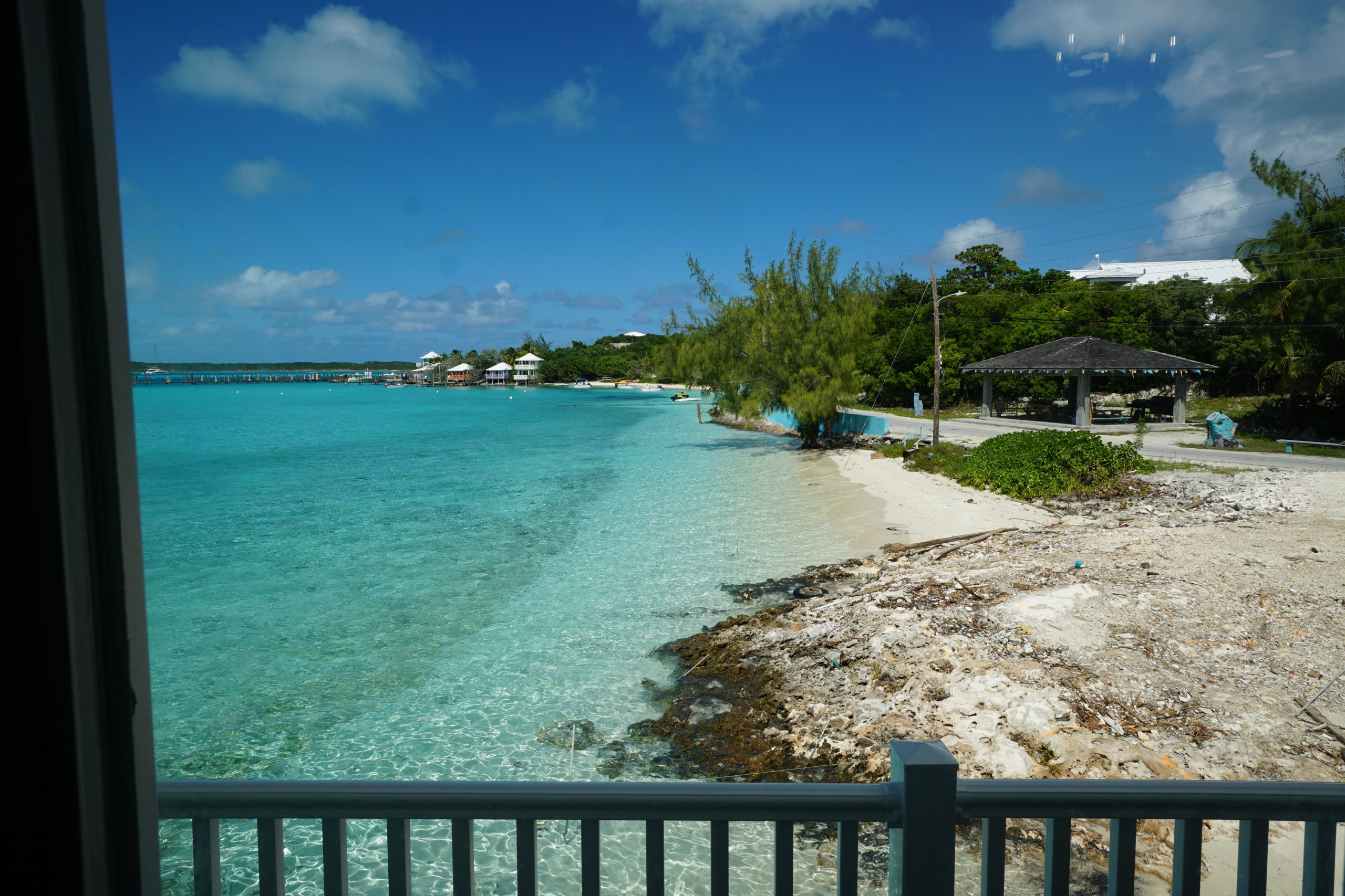 staniel Cay vacation Rentals View of a clear turquoise shoreline with a small beach, trees, and a pavilion on the right under a blue sky with scattered clouds.