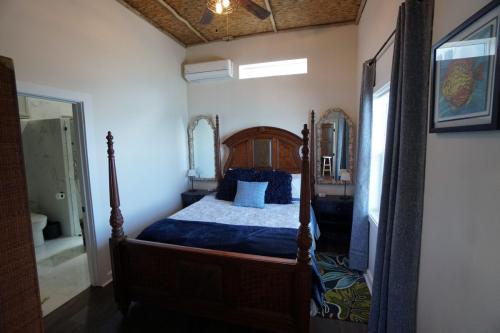 Staniel Cay Vacation Rentals, One bedroom on the water with view of the Staniel Cay Yacht Club. Bedroom with a wooden canopy bed, decorative mirrors on both sides, and a view into a bathroom. The room has white walls, a ceiling fan, and curtains on the window.