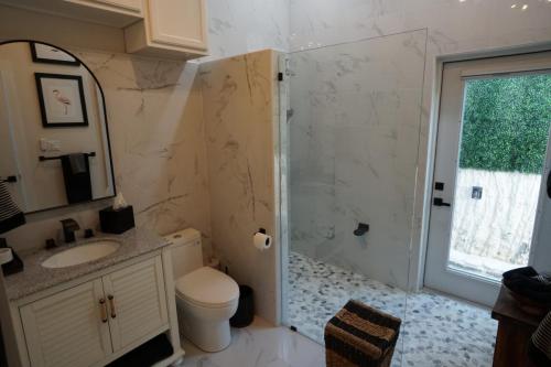 Modern bathroom with marble walls, glass shower, vanity with mirror, toilet, and a basket. A framed picture and a towel hang on the wall. Natural light from a door with a window.