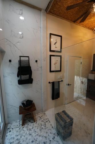 A modern bathroom with marble walls, a pebble-tiled floor, framed bird prints, and a glass shower enclosure. A wooden stool holds towels.