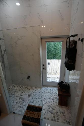 Modern bathroom with a walk-in shower featuring pebble flooring. A glass door leads outside. Towels hang on a wall rack, and a small wooden bench is placed in the corner.