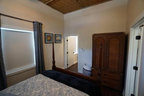 A bedroom with a four-poster bed, a wooden wardrobe, two framed pictures, and curtains covering a window. The room has a white wall and a door leading to a hallway.