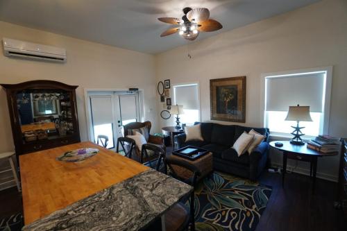 A cozy living room with a sofa, armchairs, and a wooden table. The room has a ceiling fan, patterned rug, and framed artwork on the wall, with natural light from the windows.