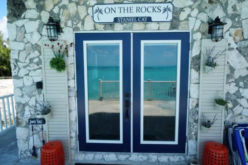 Stone building entrance with double glass doors, a sign saying On The Rocks, and views of a turquoise sea. Two lanterns and plants are beside the door. A small sign reads Staniel Cay.