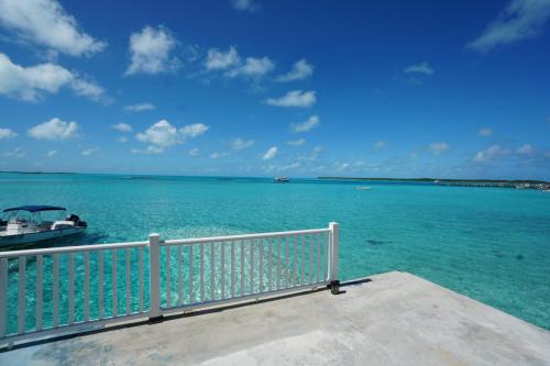 Clear turquoise ocean with a few boats, viewed from a concrete pier with a white railing. Blue sky with scattered clouds above. Staniel Cay Vacation Rentals, One bedroom on the water with view of the Staniel Cay Yacht Club.