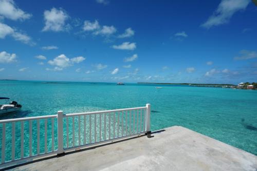 Staniel Cay Vacation Rentals, One bedroom on the water with view of the Staniel Cay Yacht Club. Clear turquoise ocean with a white fence in the foreground and a blue sky with scattered clouds in the background.