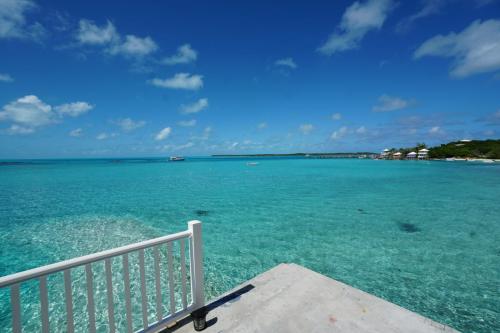 Staniel Cay Vacation Rentals, One bedroom on the water with view of the Staniel Cay Yacht Club. A clear turquoise ocean under a blue sky with fluffy clouds. White railing and concrete platform in the foreground. Distant shoreline with small buildings and greenery.