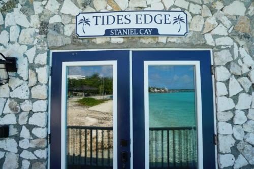 Glass doors reflecting a beach scene with blue water and a clear sky, under a sign reading Tides Edge, Staniel Cay on a stone wall.