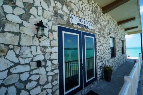 Stone building with blue double doors and a sign reading Tides Edge. A wall lantern is on the left, and the ocean is visible in the background.