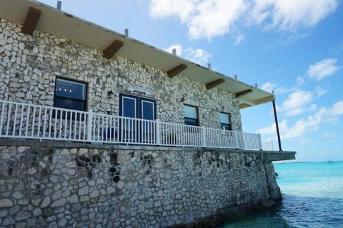 A stone building with a wraparound balcony overlooks a clear blue ocean under a partly cloudy sky.