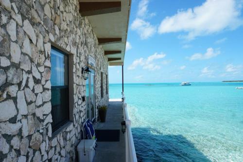 Stone building with a balcony overlooking clear turquoise ocean water under a blue sky with scattered clouds. A boat is visible in the distance.