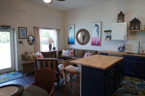 A cozy living room and kitchen area with a sofa, coffee table, wall art, and bar-style island. The room has light walls, a window, and a mix of wicker and upholstered furniture.