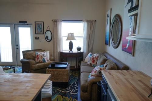 Living room with wicker furniture, colorful cushions, a wooden table, two windows, paintings on the walls, and a lamp on a side table.