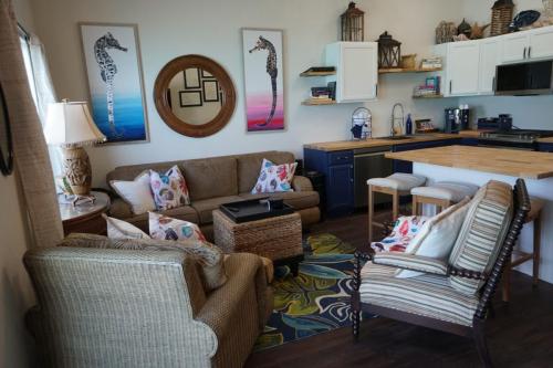 Living room with wicker and upholstered seating, a round mirror, seahorse art, and a small open kitchen with wooden countertops.