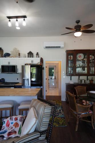 A cozy living space with a ceiling fan, kitchen counter with stools, appliances, wooden cabinet, and open doorway. Colorful throw on a striped chair in the foreground.