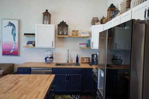 A modern kitchen with navy and wooden cabinetry, a stainless steel fridge, and various decorative items including lanterns and books on shelves. A seahorse painting adorns the wall.