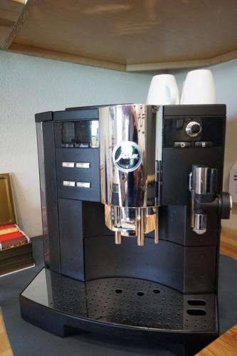 A black espresso machine with chrome accents is placed on a counter. Two white cups are on top, and a small box of tea sits to the left.