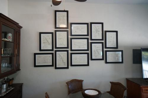 A wall with eleven framed blueprints arranged in a collage above a dining table and chairs. Nearby is a wooden cabinet and part of a dark kitchen countertop.