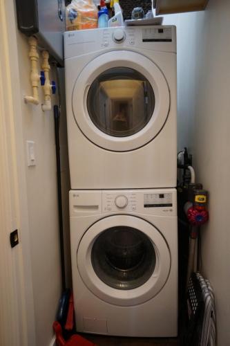 Stacked white washer and dryer units in a narrow laundry area with cleaning supplies and tools placed around them.