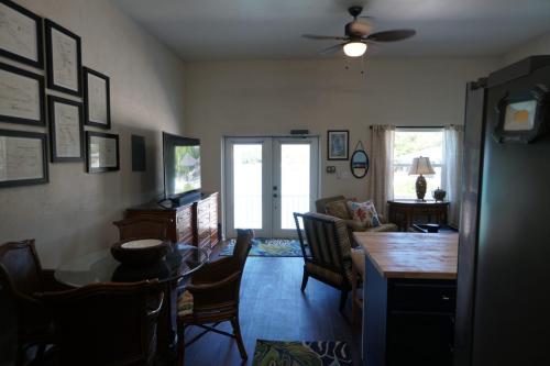 Cozy living area with a ceiling fan, framed artwork on the wall, a TV, and wicker furniture. A door with glass panels leads to an outdoor area. Light filters through a small window on the right.