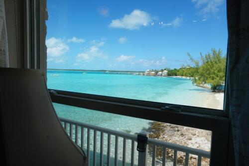 View from a window showing a clear turquoise sea, sandy beach, and distant buildings under a blue sky with scattered clouds.