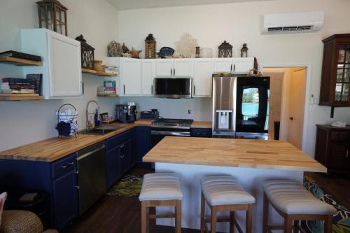 Modern kitchen with dark blue and white cabinets, wooden countertops, stainless steel appliances, and an island with bar stools. Shelves hold decorative items and books.