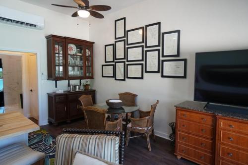 A cozy living room features wicker chairs around a glass table, framed documents on the wall, a TV on a dresser, and a ceiling fan.