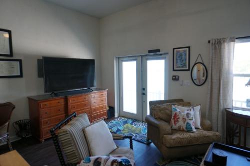 A living room with a flat-screen TV on a wooden dresser, chairs with patterned cushions, a rug on the floor, and French doors with curtains.