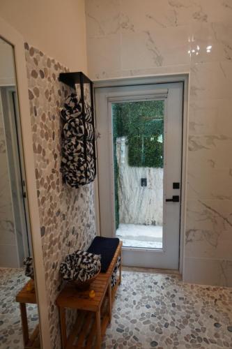 Modern bathroom with pebble-tiled walls and floor, a wooden bench with folded towels, and a glass door leading to an outdoor area.