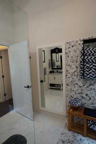 A bathroom with a large mirror reflecting a vanity. Pebble-tiled wall, wooden bench, and navy patterned towels are visible. White door is ajar in the background.