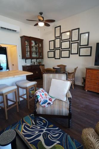 Vacation On Staniel Cay, BahamasA cozy living room features a striped armchair with cushions, a patterned rug, a ceiling fan, framed art on the wall, a wooden cabinet, a dining table with chairs, and a kitchen counter with stools.