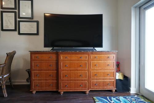 Vacation On Staniel Cay, BahamasA flat-screen TV sits on a wooden dresser with multiple drawers, next to a wicker chair and a patterned rug, beneath framed art on the wall.