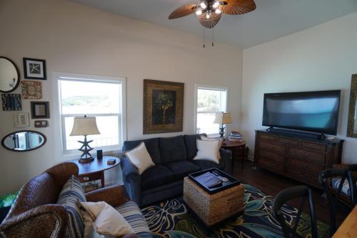 Staniel Cay Vacation Rentals, One bedroom on the water with view of the Staniel Cay Yacht Club. A cozy living room with a sofa, two chairs, a large TV on a wooden stand, and decorative elements, including lamps, wall art, and a patterned rug, under a ceiling fan.