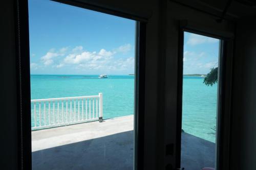 Staniel Cay Vacation Rentals, One bedroom on the water with view of the Staniel Cay Yacht Club. View through open doors revealing a turquoise ocean, white railing, and a distant boat under a blue sky with scattered clouds.